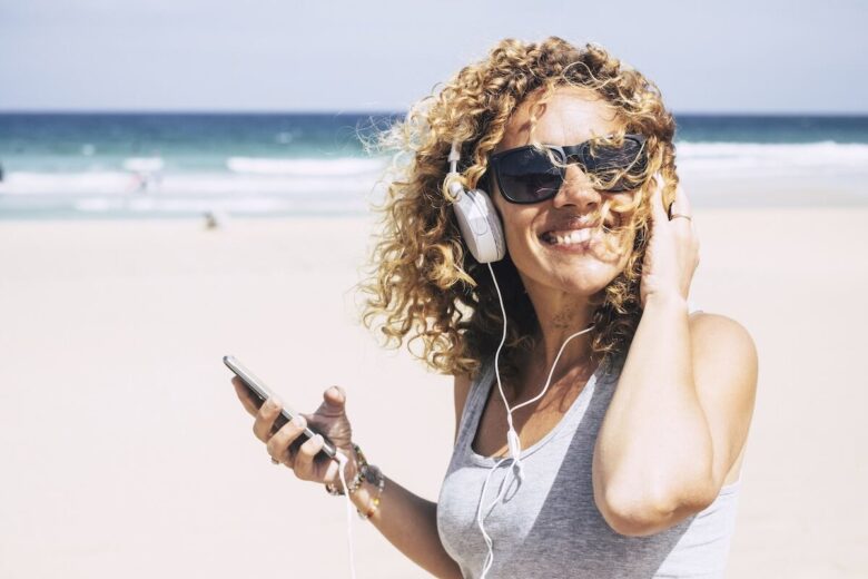 Donna bionda sorridente su una spiaggia con smartphone in mano e cuffie per ascoltare musica sulle orecchie