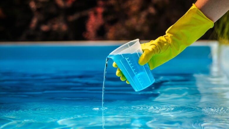 Mano con guanto giallo che versa un bicchiere di cloro-disinfettante in una piscina