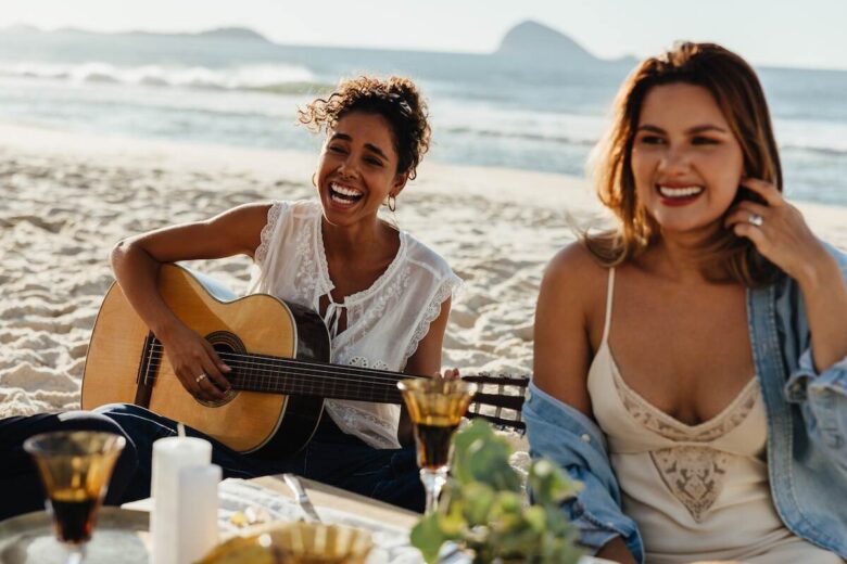 Due ragazze su una spiaggia, una suona la chitarra e canta, l'altra sorride