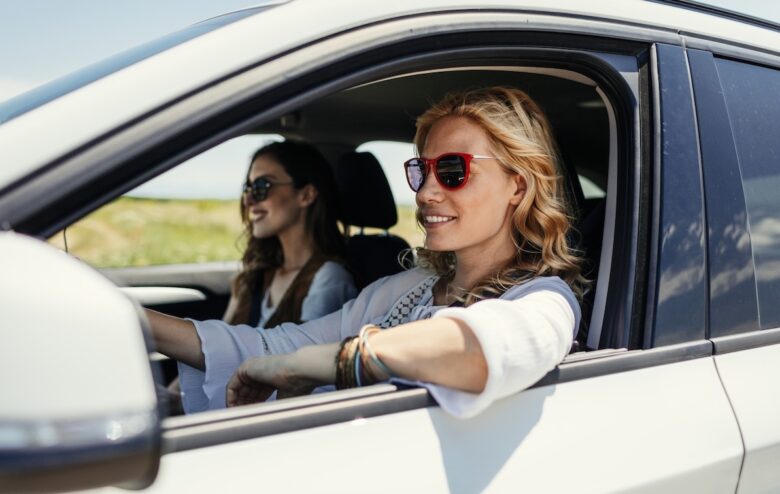 Donna bionda e sorridente al volante di una macchina bianca con accanto un'amica