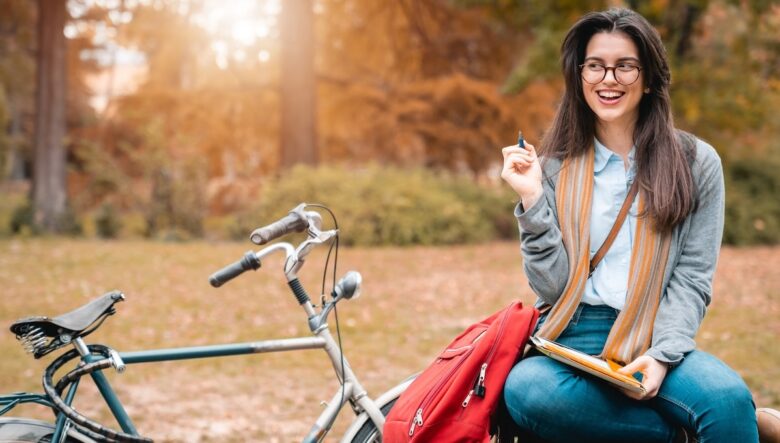 ragazza sorridente con in mano un libro-quaderno e una penna e, accanto, una bicicletta