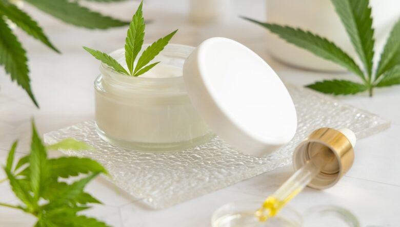 Barattolo di crema bianca aperto con un coperchio vicino alle foglie di cannabis