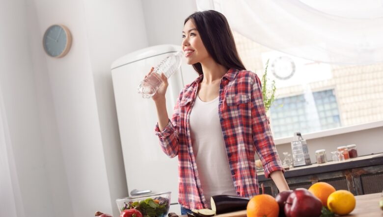 Ragazza mora fotografata in una cucina che beve acqua da una bottiglietta; davanti a lei, un tavolo con frutta e verdura