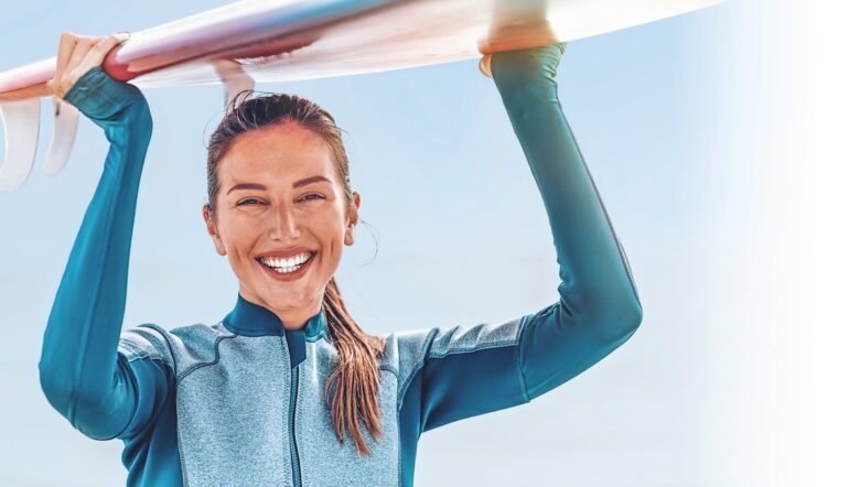 Ragazza con la muta tiene una tavola da surf sopra la testa e sorride